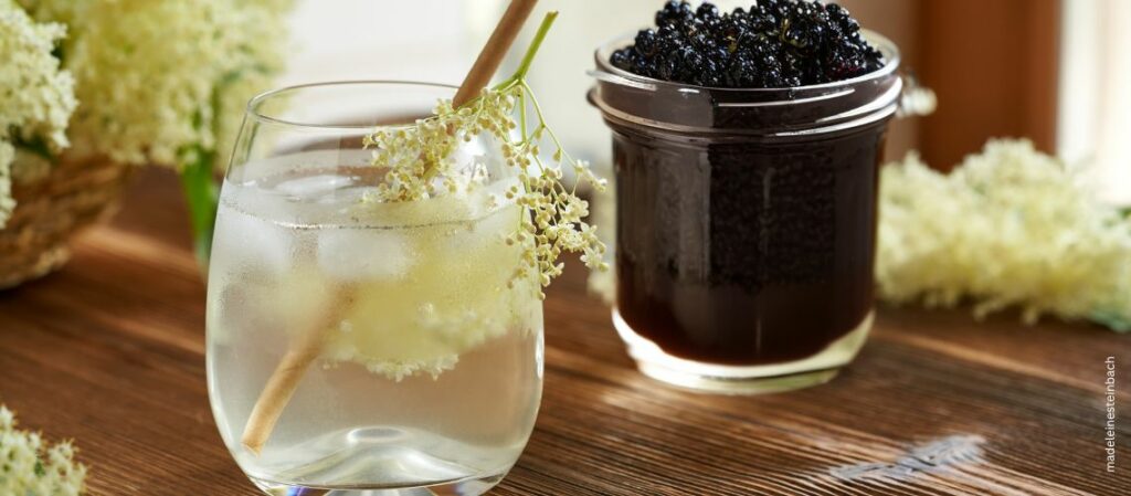 gekühltes Getränk mit Holunderblütensirup und frischen Holunderblüten. Im Hintergrund ein Glas mit Holunderbeerengelee und große Dolden mit frischen Holunderblüten zum Thema Das sind Omas beste Holunderrezepte