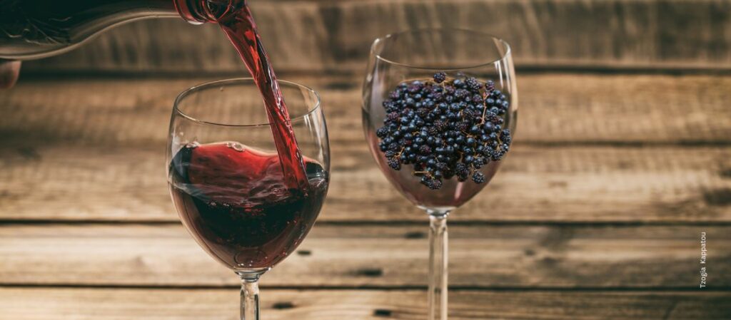 Ein Blas Rotwein, bei dem gerade nachgeschenkt wir und eine Glas mit Holunderbeeren zum Thema Ist Rotwein gesund! Darum ist Holunder gesünder